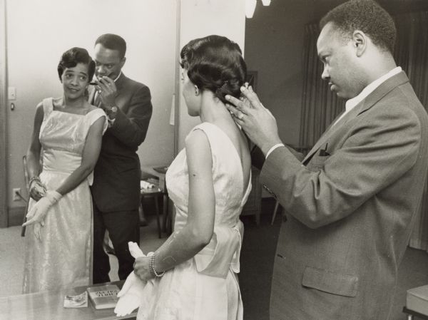 Vel Phillips standing in front of a mirror. She is wearing a white dress and gloves. There is an unidentified man behind her wearing a suit. He is holding a comb and helping Vel with her hair.