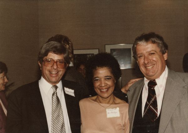Group portrait of Wisconsin Secretary of State Vel Phillips wearing a pink top and a necklace standing between two men. On the left is Wisconsin Attorney General Bronson La Follette, and on the right is Wisconsin Treasurer Charles P. Smith.