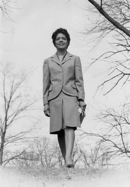Full-length portrait of Vel Phillips wearing a suit and skirt, and holding a pair of gloves in her left hand. She is walking outside on grass with trees in the background.