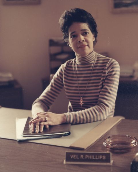Vel Phillips sits behind a desk wearing a striped turtleneck and a necklace. She is resting her hands on a folder on the desk blotter. Her nameplate is at the front of the desk near an ashtray.