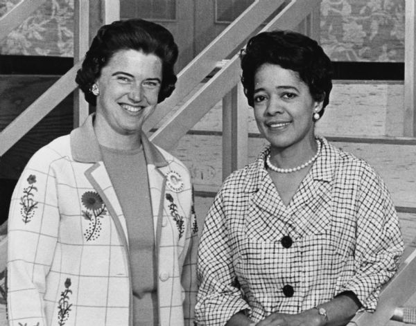 Vel Phillips, standing on the right, is wearing a necklace and earrings and a checked blouse. On the left is Carol Baumann who is wearing a floral jacket and a button pin with her portrait that reads: "Baumann Ninth Dist. Congress." Baumann ran for Wisconsin Congress from the ninth district.
