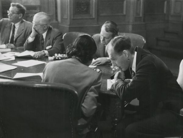 View over back of chair towards Vel Phillips sitting at a table talking with an unidentified man on her right. In the background, sitting on the other side of the table, is Alfred C. Hass (second from left).