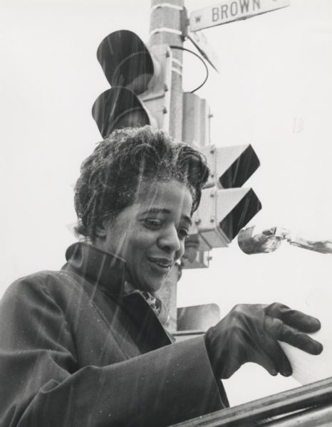 View looking up towards Vel Phillips standing at a podium. She is wearing a coat and gloves and speaking into a microphone. Snow is falling. There is a traffic light signal above and behind Vel, with a street sign that reads: "W Brown Street."