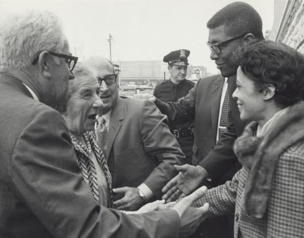 Vel Phillips, wearing a coat and fur scarf, is shaking the hand of Golda Meier. There are other unidentified men surrounding the two. In the background is a police officer.