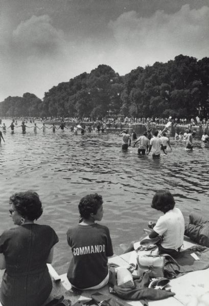 Rear view of Vel Phillips sitting on a pier on a lake. She is wearing a shirt that says "Commando" on the back. Two unidentified women are sitting next to her. In the water are protesters, a number of them holding hands in a line. A large group of people are along the shoreline in the background.