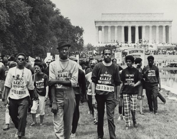 Vel Phillips (left) is marching with the Milwaukee NAACP Youth Council. In the background is the Lincoln Memorial. Other unidentified youth council members are marching. One person is wearing a shirt that says "I'm tired of drinking from my ghetto cup." There are other protest signs in the background.