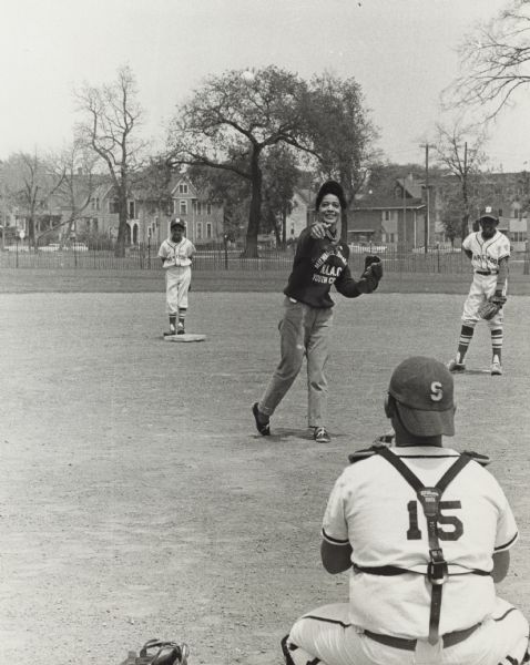 View from behind the catcher towards Vel Phillips throwing a baseball pitch. She is wearing a sweatshirt that reads "Milwaukee Commandos NAACP Youth Council." The catcher and two other boys are wearing Shorewood baseball uniforms. There is a fence and houses in the background.