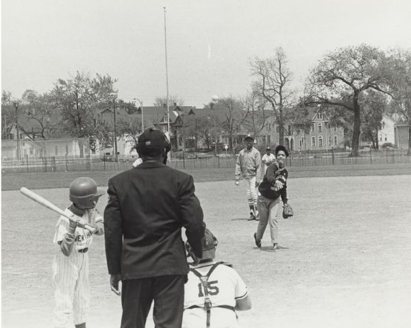 Vel Phillips, wearing an Milwaukee Commandos NAACP Youth Council sweatshirt, throws a pitch. A batter, catcher, and umpire are in the foreground. Two unidentified children are behind Vel. There is a fence and houses in the background.