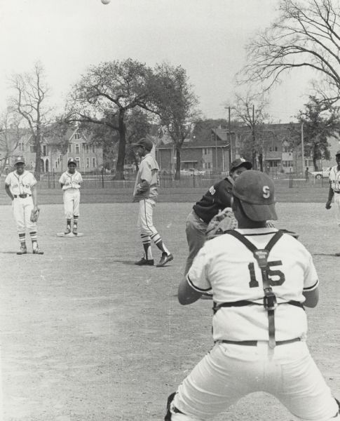 View from behind catcher towards Vel Phillips, wearing an Milwaukee Commandos NAACP Youth Council sweatshirt, throwing a pitch to the catcher, who is standing. Four unidentified you are standing behind Vel. Their uniforms say "Shorewood." There is a fence and houses in the background.