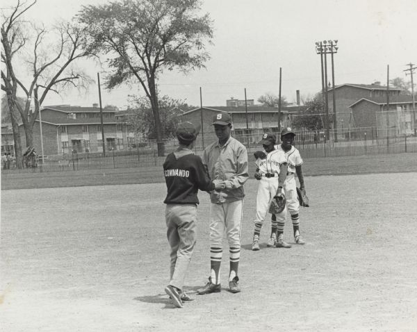 Vel Phillips, wearing an Milwaukee Commandos NAACP Youth Council sweatshirt, handing a baseball to an unidentified young man wearing a jacket. Two unidentified children are behind them wearing uniforms that read "Shorewood." There is a fence and houses in the background.