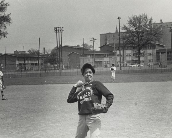 Vel Phillips, wearing an Milwaukee Commandos NAACP Youth Council sweatshirt, throwing a baseball. Two unidentified children stand in the field behind Vel. There is a fence and houses in the far background.