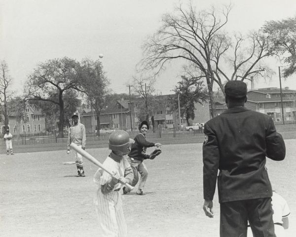 View from behind batter and umpire of Vel Phillips pitching a baseball. She is wearing a Milwaukee NAACP Youth Council sweatshirt. There are three unidentified children wearing baseball uniforms behind her. In the background is a fence, houses, and trees.