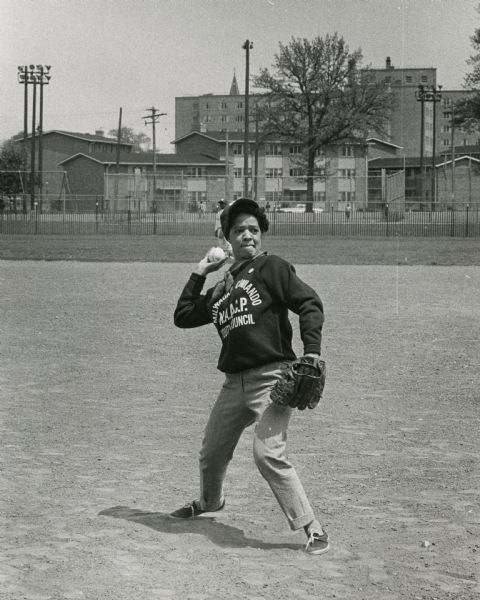Vel Phillips, wearing a baseball glove, hat and a sweatshirt that reads "Milwaukee Commando NAACP Youth Council" pitching a baseball. In the background standing in the field behind her is an unidentified child wearing a baseball uniform. In the far background are trees, houses, and a fence.