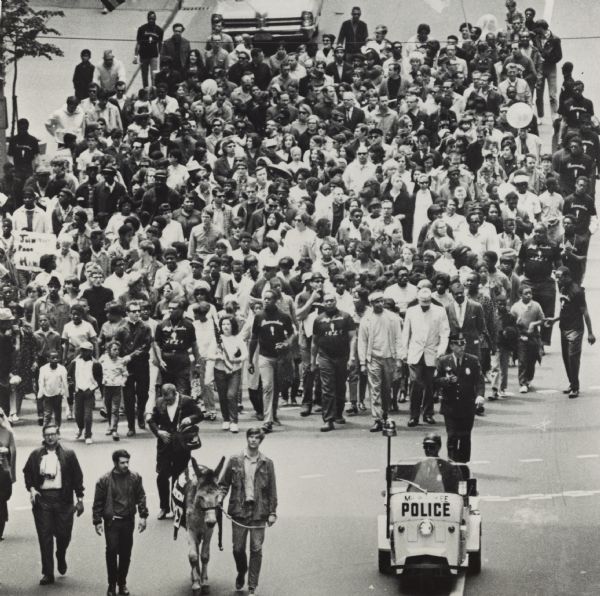 Elevated view of a group of people marching in the street with Father James Groppi. There is a man driving a Milwaukee police vehicle in front, and one man is leading a donkey, which is wearing a banner over its back.