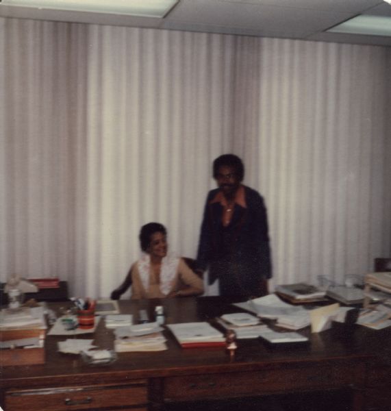 Vel Phillips sitting behind the large Wisconsin Secretary of State desk. An unidentified man is standing next to her.