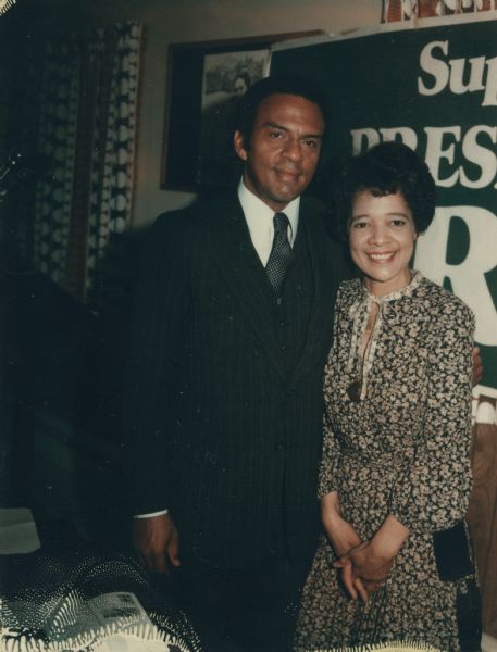 Vel Phillips, in a flower patterned dress, standing next to an unidentified man who is wearing a suit and necktie. Behind them is a sign that partially reads: "Sup___ Pres___."