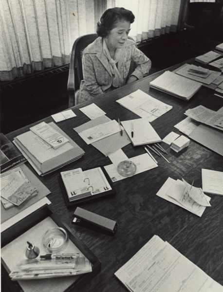 View across desk filled with papers towards Vel Phillips sitting while serving as Wisconsin's Secretary of State.