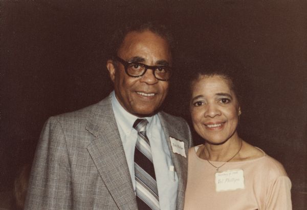 Quarter-length portrait of Vel Phillips, wearing a name tag that reads "Secretary of State," standing next to her husband, W. Dale Phillips, who is wearing a suit and eyeglasses.