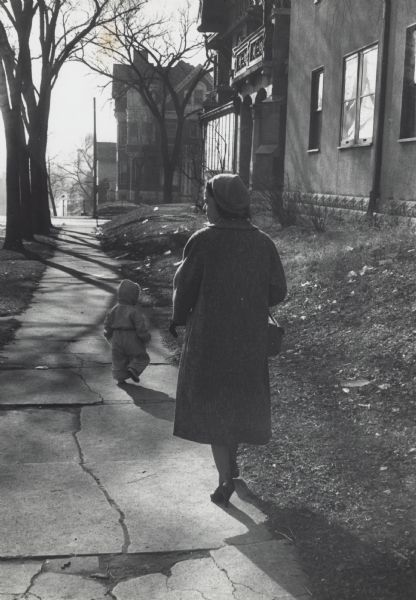 Rear view of Vel Phillips walking on a sidewalk in a neighborhood, with a small child walking in front of her. Vel is wearing a long coat and a hat.