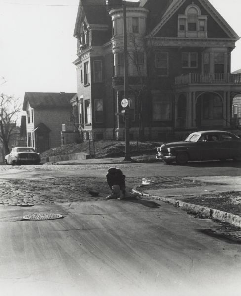 View down street towards Vel Phillips, who is crouching down near the curb at an intersection helping a child laying in the street. Her handbag is next to her. There are two automobiles in the background. The street signs read 5th Street and Galena Street.