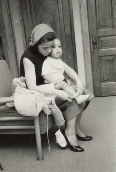 Vel Phillips sitting in a chair with her son, Dale Phillips, in her lap. She is helping him put on shoes and a coat.