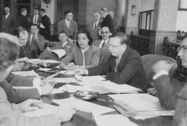 View across table towards Vel Phillips seated with other unidentified men, possibly other common council members. Sheets of paper are scattered on the table. The group appears to be in the Milwaukee Common Council chamber.
