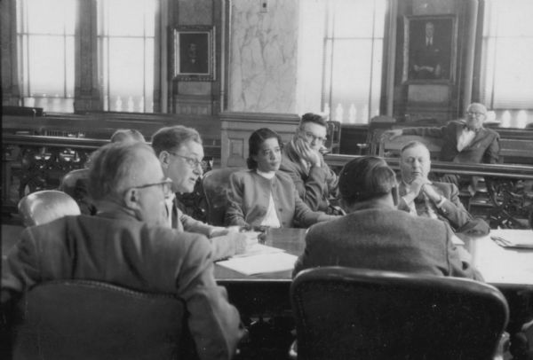 View across room towards Vel Phillips sitting at a table in the Milwaukee Common Council chambers with a group of unidentified men. There are papers on the table.