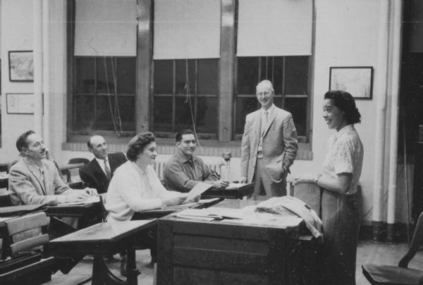 View from side of classroom towards Vel Phillips who is standing behind a teacher's desk. There are a group of unidentified adults sitting in the classroom seats on the left. An unidentified man is standing in the background.
