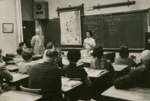 View from back of classroom towards Vel Phillips standing behind a desk. The chalkboard behind her reads: "Protective" and "Public Works." There is a map of Milwaukee hanging above the chalkboard on the left. A group of men and women are sitting in the desks listening, and a man stands at the front of the classroom on the left.