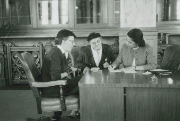 Vel Phillips sitting at her desk in the Milwaukee Common Council chamber. She is writing on a piece of paper. Seated next to her are two unidentified women wearing hats.