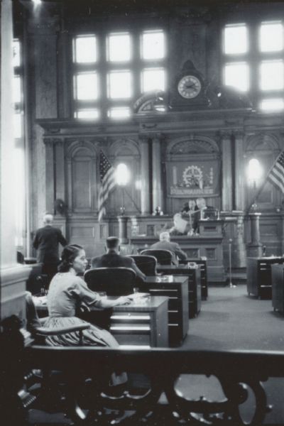 Rear view of Vel Phillips sitting at her desk in the Milwaukee Common Council chamber. Other unidentified council members are sitting at their desks. The main speaker podium is in the background with two American flags and a banner that reads "Milwaukee."