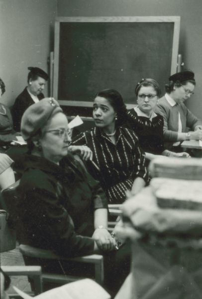 Vel Phillips, wearing a striped dress, is sitting at a table with a group of unidentified women. There is a chalkboard in the background.