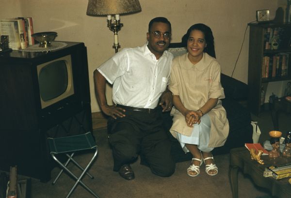 Vel Phillips sitting in an armchair, wearing a tan top with a light blue skirt. Dale is kneeling next to her, wearing eyeglasses. A television is in the corner and a lamp is in the background.