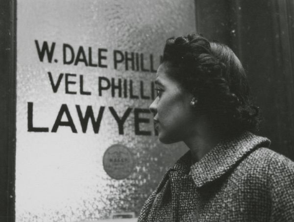 Vel Phillips standing in front of the door to her and her husband's law office. The sign on the glass door reads: "W. Dale Phillips, Vel Phillips Lawyers." There is a NAACP sticker below the names.