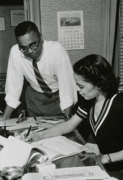 Vel Phillips sitting at a desk writing on a legal pad. Looking on is her husband W. Dale Phillips.