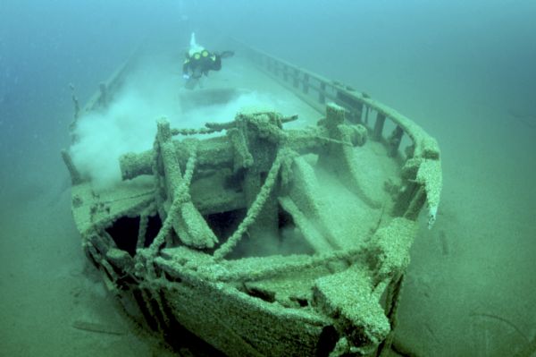 Underwater view of the starboard side of the bow of the schooner <i>Home</i>, the site of the collision damage that sank the ship. The sampson post and windlass are sitting in the foreground, with ropes hanging off from the windlass over the starboard side of the ship. A scuba diver (an underwater archeologist) is swimming over the deck of the <i>Home</i> just behind the collision damage.