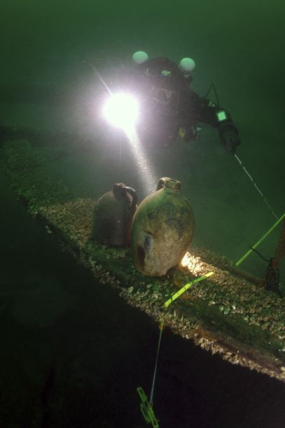 Underwater view of a scuba diver (an underwater archaeologist) shining a light on two liquor or water jugs that had been pulled out during the archaeological survey for measuring. The vessels are sitting on part of the wreckage of the sunken schooner <i>Home</i>. The diver has also placed a measuring tape across the mussel covered wood on which the vessels are sitting.