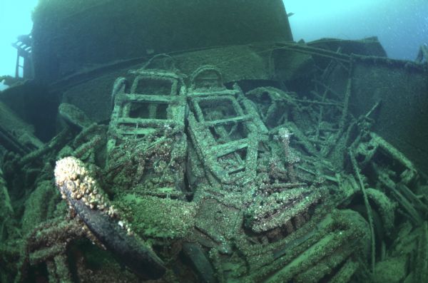 Underwater view of metal frames and rubber wheels of automobiles laying in a pile on top of each other within the sunken <i>Lakeland</i>. Part of the upper deck of the <i>Lakeland</i> can be seen behind its cargo of cars.