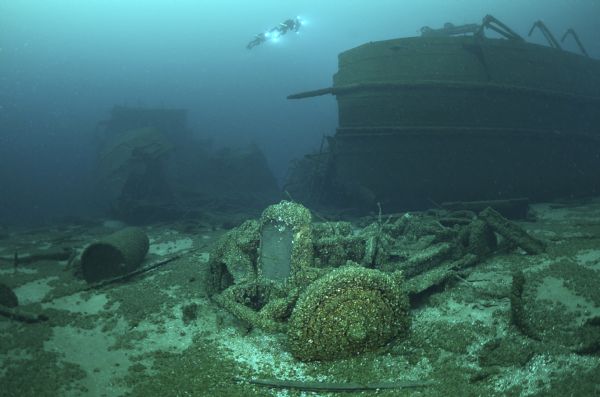 Underwater starboard view of the <i>Lakeland</i>. While the ship remains largely intact, it is split in half, with the back end of the ship in the background, and the front half visible along the right side. Two underwater archeologists swim over the <i>Lakeland</i>. In the foreground an automobile is sitting on the lake floor, a part of the ship's lost cargo.