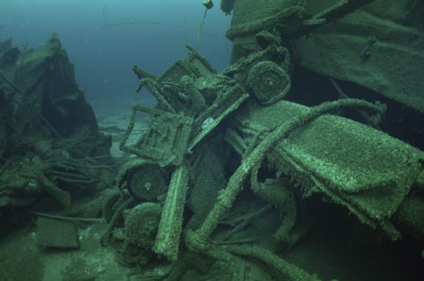 Underwater view of an automobile hanging off of the ruins of the steamer <i>Lakeland</i>. The wheels are still attached to the car, but most of the outward frame is gone, with the door hanging on its hinges. Other car parts and pieces of the sunken <i>Lakeland</i> are laying on the floor of Lake Michigan.