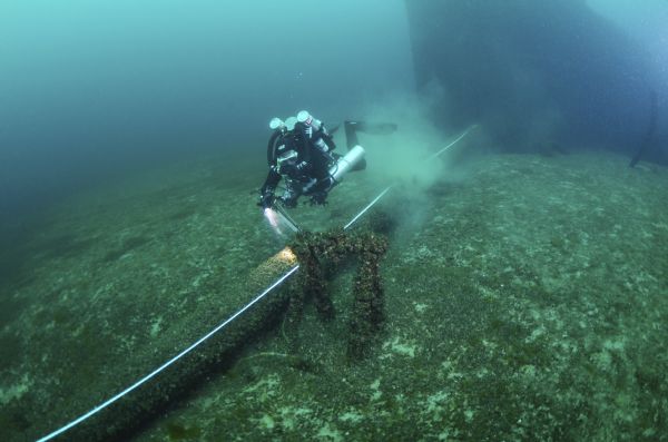 Underwater view of an archeologist surveying the site of the sunken <i>Milwaukee</i>. The archeologist is swimming over a measuring tape, which runs from the ship (in the background) to a site leading off to the left.