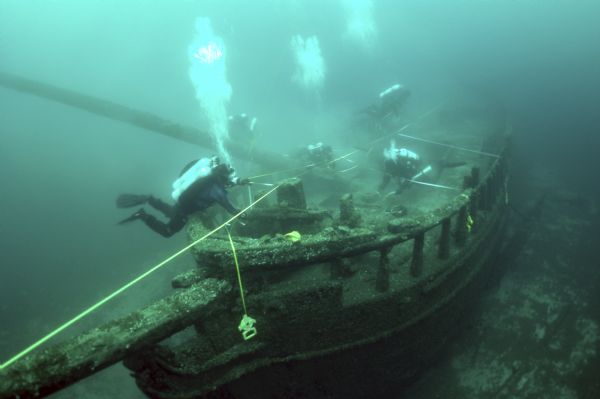 Underwater view of five maritime archeologists measuring and surveying the bow of the sunken <i>Northerner</i>. Much of the ship appears intact, except the foremast which is bent over the starboard side.