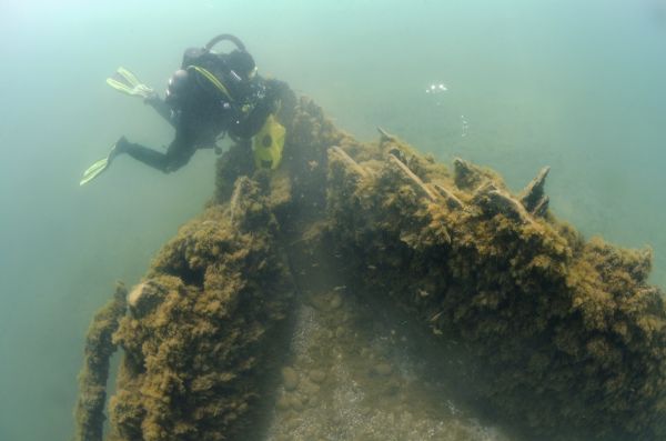 Underwater view of the bow of the <i>Pathfinder</i>. Algae is growing along the sides, and clumps of sand are on the ship. A maritime archeologist is surveying the bow.