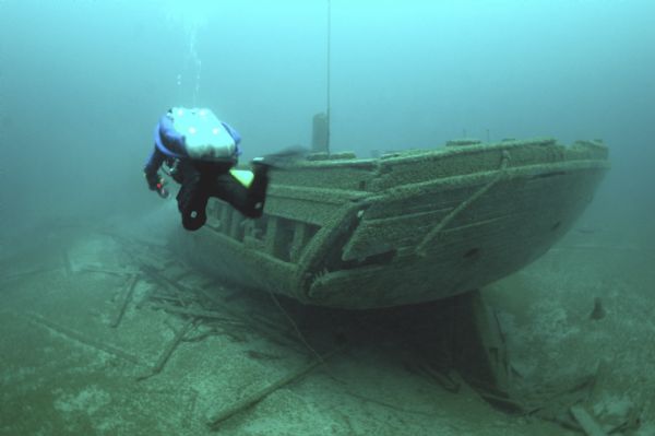 Underwater view of an archaeologist swimming past the stern of the <i>Rouse Simmons</i> (also known as the Christmas tree ship). Except for the masts, the schooner remains largely intact. Debris surrounds the ship on the lake bed.  