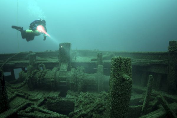 Underwater view of an archaeologist swimming over the windlass of the <i>Rouse Simmons</i> (also known as the Christmas Tree Ship). The archaeologist is shining a light on the windlass. Chains are running from the windlass and are sitting on the deck. Some planks of wood from the deck are lying bent or broken, with few sticking straight up.