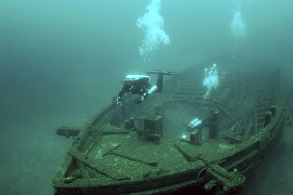 Underwater view of two archaeologists surveying and investigating the bow of the <i>Rouse Simmons</i> (also known as the Christmas Tree ship). One person is swimming directly over the bow, while the other person explores the hull.