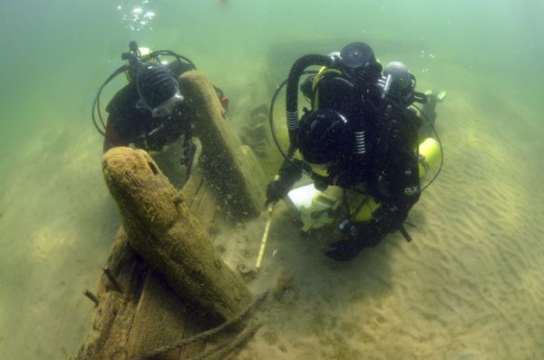 Underwater view of two archaeologists measuring and surveying the site of the <i>Success</i>. Much of the ship remains buried under sand.