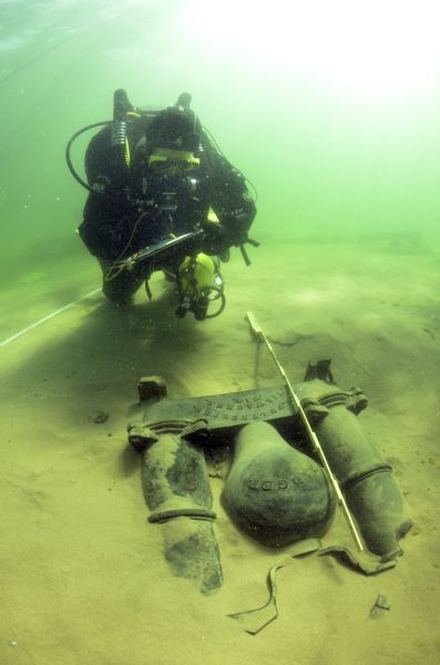 Underwater view of an archaeologist surveying the bilge pump of the <i>Success</i>. The pump is resting in a bed of sand towards the stern of the ship. Text on the pump reads: "Badger" and "Felt Housen, Milwaukee, Wis."