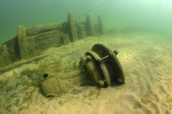 Underwater view of the capstan on the scow schooner <i>Success</i>. It is laying on its side partially buried in sand. A section of the starboard side of the <i>Success</i> is sticking out of the sand behind the capstan. 