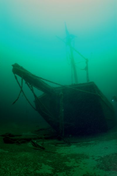 Underwater view of the wrecked scow schooner <i>Silver Lake</i>. The ship remains largely intact, but it is split in half where it was run over by the <i>Pere Marquette</i> prior to its sinking. An anchor is resting on the sand forward of its bow ramp. The ship’s foremast remains standing and rigged with a yard. 